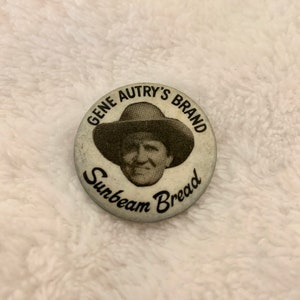 Puede incluir: Un botón blanco con una imagen en blanco y negro de Gene Autry con un sombrero de vaquero. El texto "Gene Autry's Brand Sunbeam Bread" está impreso alrededor de la imagen.