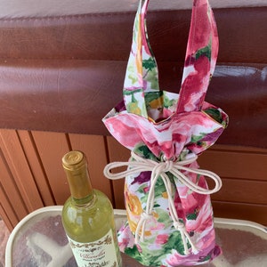 Op de afbeelding: Een bloemen wijnfles tas met een wit trekkoord en een handvat. De tas is gemaakt van stof met een roze, groene en gele bloemenprint. Een fles witte wijn staat op een tafel naast de tas.