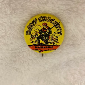 Op de afbeelding: Een gele knop met een cartoonillustratie van een man met een geweer en de tekst "Davy Crockett" en "Sunbeam Bread".