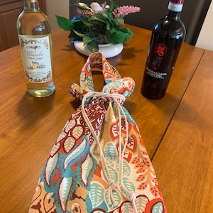 Op de afbeelding: Een kleurrijke wijnzak van stof met een trekkoord. De zak is gevuld met een fles witte wijn en een fles rode wijn. De fles witte wijn heeft een etiket met de tekst "Villanella Pinot Grigio Alto Adige". De fles rode wijn heeft een etiket met de tekst "Montepulciano d'Abruzzo".
