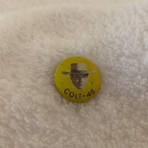 May include: A yellow button with a black and white image of a man wearing a cowboy hat. The text "COLT-45" is printed below the image.