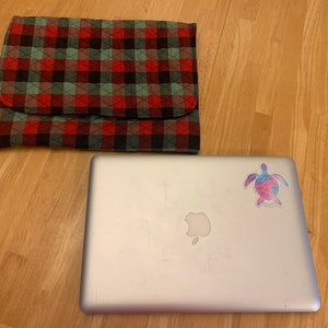 May include: A silver laptop computer with a colorful turtle sticker on the lid. The laptop is sitting on a wooden surface next to a red and green plaid laptop sleeve.