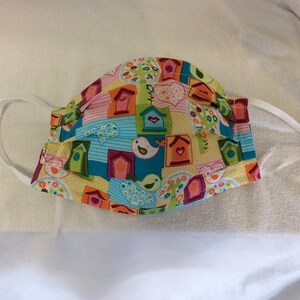 May include: A yellow, blue, green, pink, and red fabric face mask with a pattern of birdhouses and birds. The mask has white elastic ear loops.