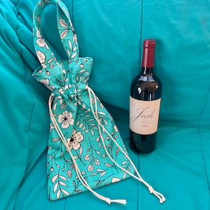 Op de afbeelding: Een turquoise stoffen wijnzak met een wit bloemenpatroon en een wit trekkoord. De zak ligt op een turquoise deken met een fles rode wijn.