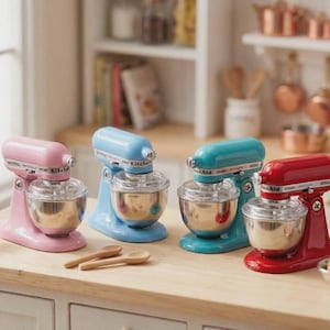 May include: Miniature stand mixers in pink, blue, teal, and red with stainless steel bowls. Wooden spoons and a small scale are on the wooden surface. A bag of flour and a bowl of eggs are also present.