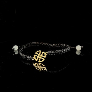 Op de afbeelding: Een zwarte macramé armband met een goudkleurig, geometrisch middelpunt. De armband heeft twee witte kralen en wordt gepresenteerd tegen een zwarte achtergrond. De armband is verstelbaar.