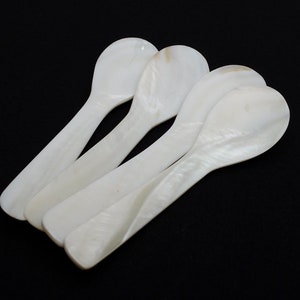 May include: Four white mother of pearl spoons, each with a rounded bowl and a long, slender handle. The spoons are arranged in a stack, with the handles overlapping.