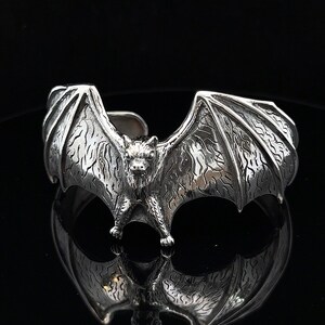 May include: A silver-colored cuff bracelet featuring a detailed bat design with outstretched wings. The bat's wings are textured, and the bracelet has an open design for easy wear. The jewelry is displayed on a reflective surface.