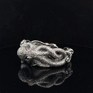 May include: A silver ring with an octopus design. The ring has a textured surface detailing the octopus's head and tentacles, with a polished finish. The ring is displayed on a reflective black surface.