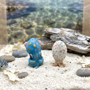 May include: Two ceramic octopus figurines. One is blue with yellow spots, the other is white with red and blue spots. They are on a bed of white sand with small rocks and a piece of driftwood in the background. The octopus figurines are small, decorative items.