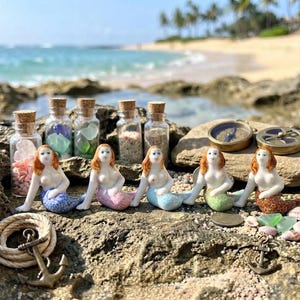 May include: Five small ceramic mermaid figurines with red hair and white skin, each featuring a unique colored tail in blue, pink, light blue, green, and brown. They are displayed with five glass bottles filled with sand and sea glass, two anchors, and a compass.