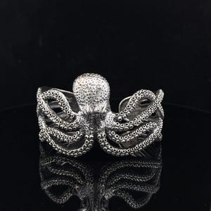 Könnte beinhalten: Ein silberfarbenes Armreif-Armband mit einem Oktopus-Motiv. Das Armband zeigt ein detailliertes Oktopus-Design mit strukturierten Saugnäpfen an den Tentakeln. Der Kopf des Oktopus ist zentriert, und die Tentakel umschließen das Handgelenk.
