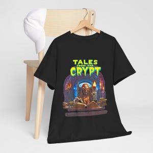 May include: A black short-sleeve t-shirt featuring a graphic print of the text Tales from the Crypt in bright green letters above an illustration of a skeletal figure sitting at a desk with an open book and a lit candle in a dark stone room.