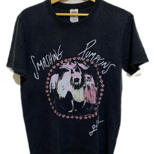 Puede incluir: Camiseta negra con la banda Smashing Pumpkins. El diseño incluye una imagen circular de los miembros de la banda y el nombre de la banda en escritura blanca. La palabra "girls" está escrita debajo.