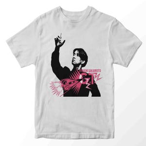Könnte beinhalten: Weißes T-Shirt mit einer schwarz-rosa Grafik einer Person, dem Text "RYUICHI SAKAMOTO" und "G.T.". Die Grafik enthält ein stilisiertes Bild einer Person, die den Arm hebt.