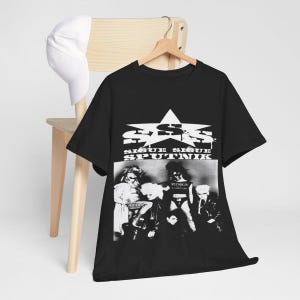 May include: Black t-shirt featuring a band photo and the text "SIQUE SIQUE SPUTNIK" in white. The shirt is hanging on a wooden hanger. A white baseball cap rests on a wooden chair.