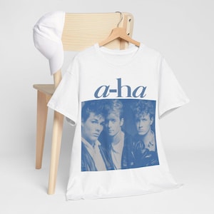 Peut inclure: T-shirt blanc avec un imprimé bleu du groupe a-ha. L'impression représente trois hommes dans un groupe, avec le nom du groupe en lettres bleues au-dessus d'eux.