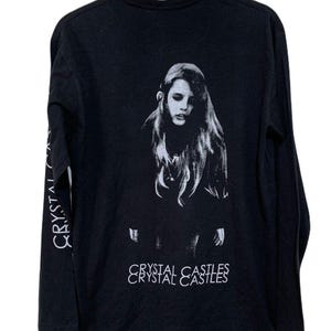 May include: Black long-sleeve shirt featuring a grayscale image of a woman on the back. The text "CRYSTAL CASTLES" is printed vertically on the left sleeve and horizontally at the bottom of the image.