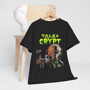 Peut inclure: Un t-shirt noir à manches courtes avec une impression graphique du Crypt Keeper et le texte "TALES FROM THE CRYPT" en lettres vert vif au-dessus du personnage. Le t-shirt est présenté sur un cintre en bois avec une casquette blanche à côté.