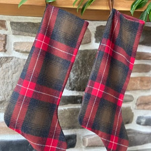 May include: Two Christmas stockings with a red, brown, and navy plaid pattern hang from a wooden mantel. The stockings are made of fabric and have a classic design. Greenery is draped above the stockings.