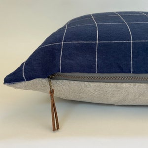 May include: A navy blue pillow with a white grid pattern and a brown leather zipper pull. The pillow has a zipper closure and a light gray linen backing.