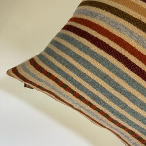 Op de afbeelding: Close-up van een gestreept kussen met een diagonaal patroon. Het kussen heeft afwisselende strepen in beige, blauw, rood, bruin en grijs. Het kussen heeft een zachte, getextureerde uitstraling, wat wijst op een geweven stof.