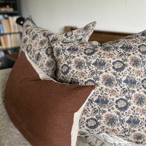 May include: Three decorative pillows on a bed. One pillow is brown with a white border. The other two pillows are white with a floral pattern in shades of blue, brown, and cream.