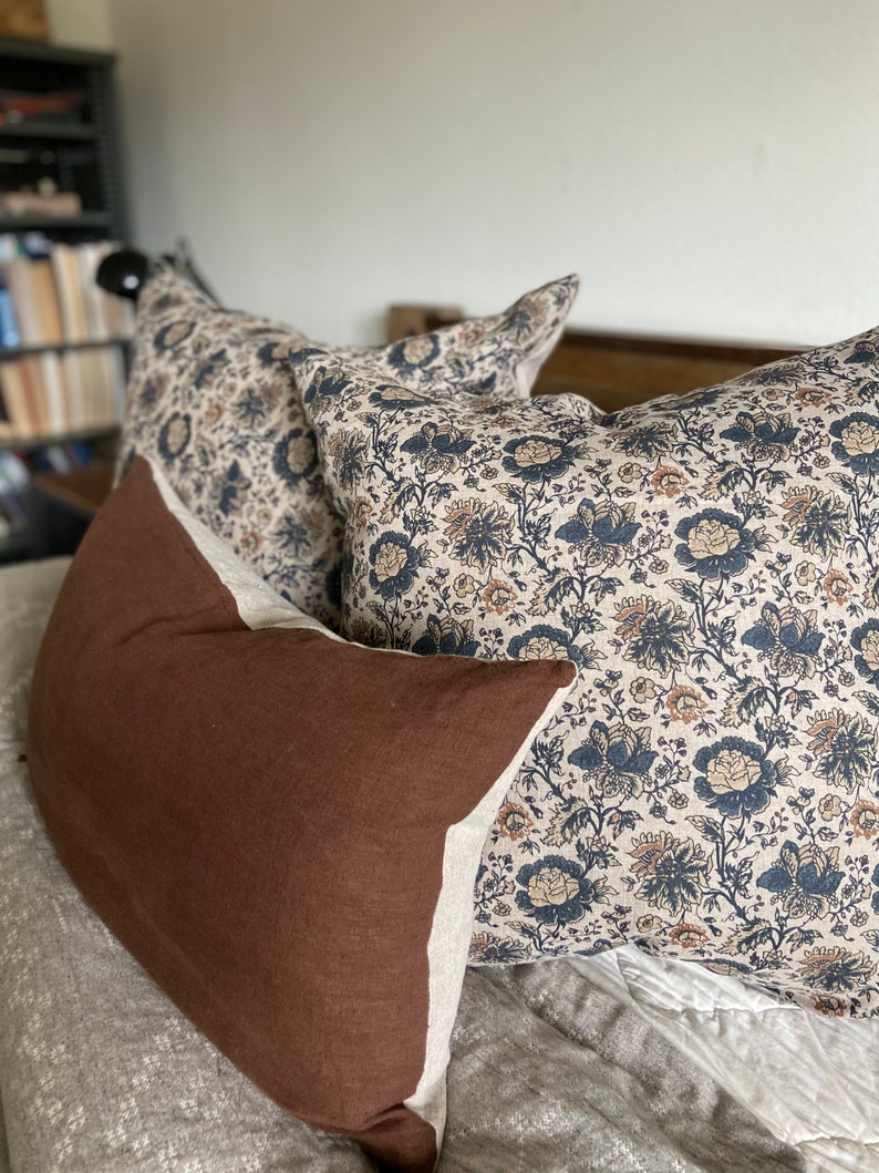 May include: Three decorative pillows on a bed. One pillow is brown with a white border. The other two pillows are white with a floral pattern in shades of blue, brown, and cream.