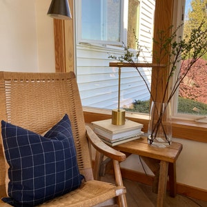 May include: A wicker rocking chair with a blue and white checkered pillow, a gold lamp with a black shade, and a wooden stool with a stack of books on top. The chair is in a room with a window and a wooden floor.