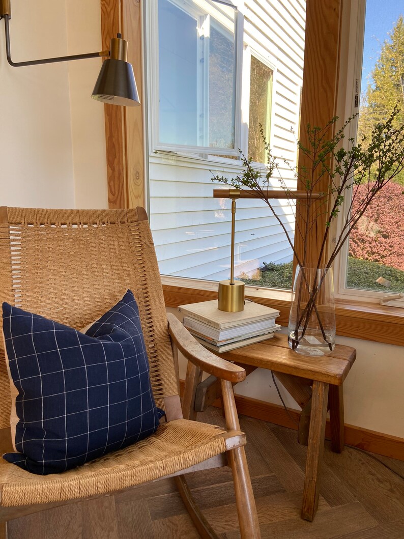 May include: A wicker rocking chair with a blue and white checkered pillow, a gold lamp with a black shade, and a wooden stool with a stack of books on top. The chair is in a room with a window and a wooden floor.