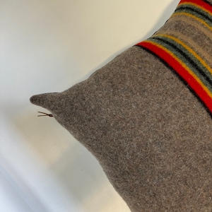 May include: A brown wool pillow with a zipper closure. The pillow has a striped pattern in red, yellow, green, and black.