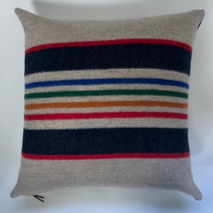 May include: A gray throw pillow with a pattern of colorful stripes in red, blue, green, orange, and brown. The pillow is made of a soft, fuzzy material.