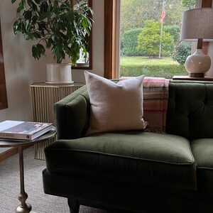 May include: A green velvet sofa with a white pillow and a red and white striped throw blanket. The sofa is in a living room with a plant, a gold side table, and a lamp.