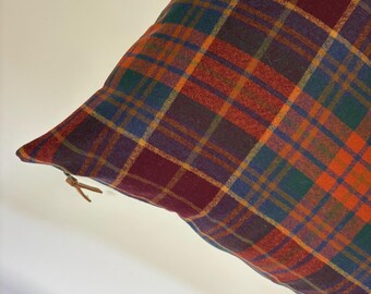 Puede incluir: Un cojín decorativo con un estampado de cuadros en tonos burdeos, naranja, verde, azul y amarillo. Una etiqueta de cuero está sujeta a la esquina. El cojín tiene forma cuadrada.