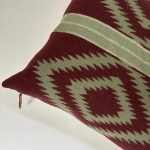 Puede incluir: Un cojín burdeos con un patrón geométrico en zigzag en verde claro. Una franja verde claro recorre la parte superior. El cojín tiene un tirador de cremallera de cuero. Este cojín decorativo es perfecto para un sofá o una cama.