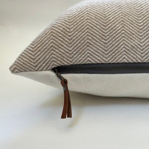 May include: A beige and white chevron patterned pillow with a zipper closure and a brown leather zipper pull.