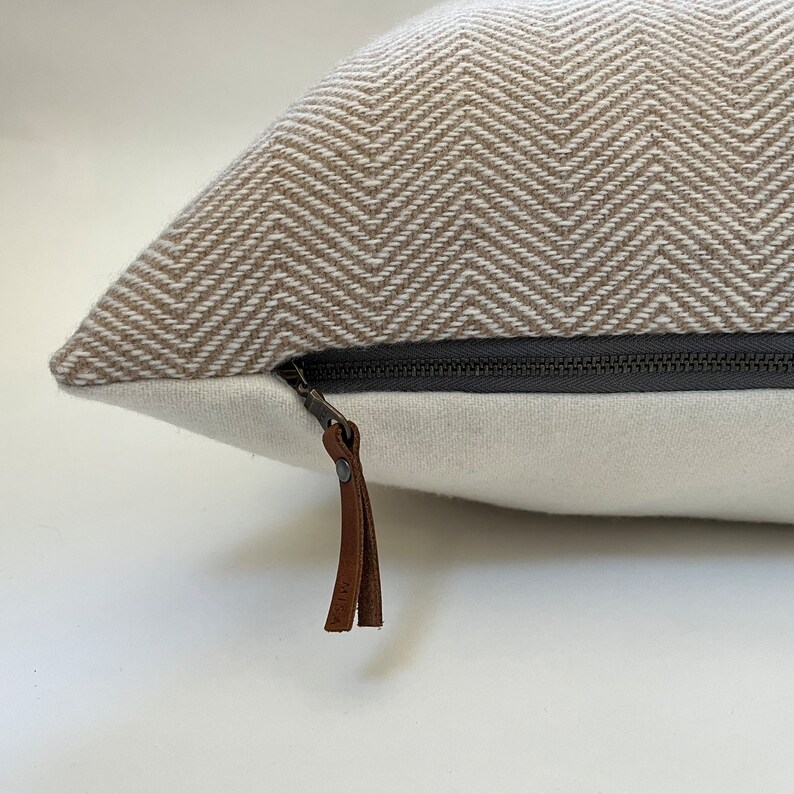 May include: A beige and white chevron patterned pillow with a zipper closure and a brown leather zipper pull.