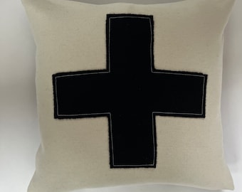 Puede incluir: Una almohada blanca con una cruz negra de fieltro cosida en la parte delantera. La cruz tiene un contorno blanco.