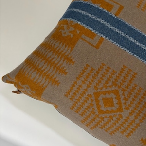 May include: A brown and tan throw pillow with a geometric pattern in shades of orange and blue. The pillow has a zipper closure and a blue stripe across the top.