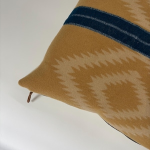 Puede incluir: Una almohada de sofá de color amarillo mostaza con un patrón geométrico y dos rayas azul oscuro. La almohada tiene una cremallera y una tira de cuero marrón.