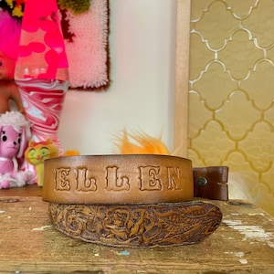 May include: A brown leather belt with the name "ELLEN" embossed on it. The belt has a floral design and is on a wooden surface.