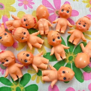 May include: A collection of vintage, peach-colored Kewpie dolls with blue eyes and black eyelashes. The dolls are lying on a floral patterned fabric with pink, yellow, and green flowers. The dolls are in various poses.