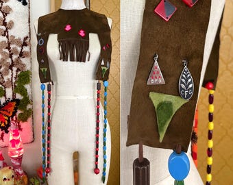 May include: A brown suede vest with fringe, beads, and colorful embroidery. The vest has two long beaded straps with red and blue beads. The vest is decorated with small, colorful geometric shapes and a green leaf design.