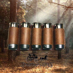 May include: A brown leather shotgun shell holder with six slots. The holder is made of brown leather and has a blue stitching detail. The holder is in front of a blurred background of trees.