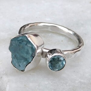 K&ouml;nnte beinhalten: Ein silberner Ring mit zwei Aquamarin-Edelsteinen. Der gr&ouml;&szlig;ere Stein ist ein roher, nat&uuml;rlicher Aquamarin, w&auml;hrend der kleinere Stein ein runder, facettierter Aquamarin ist.