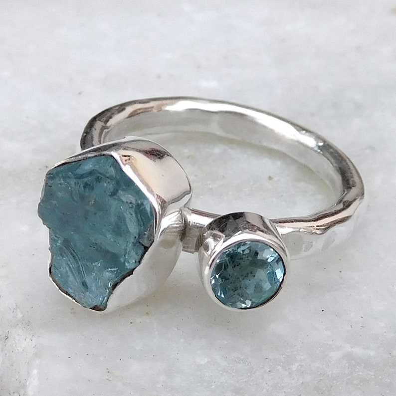 K&ouml;nnte beinhalten: Ein silberner Ring mit zwei Aquamarin-Edelsteinen. Der gr&ouml;&szlig;ere Stein ist ein roher, nat&uuml;rlicher Aquamarin, w&auml;hrend der kleinere Stein ein runder, facettierter Aquamarin ist.