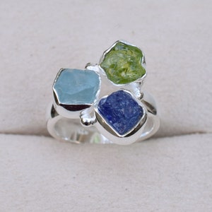 Op de afbeelding: Een zilveren ring met drie ruwe edelstenen: een lichtblauwe aquamarijn, een levendige groene peridot en een diepblauwe tanzaniet. De edelstenen zijn gezet in individuele zilveren zettingen, waardoor een clusterontwerp ontstaat.