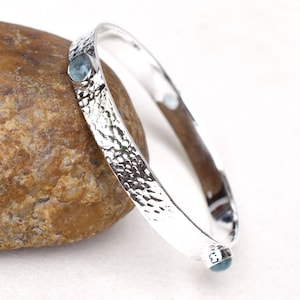 Può includere: Un braccialetto a cerchio d'argento con una texture martellata e due piccole gemme rotonde azzurre. Il braccialetto è esposto su una roccia marrone chiaro su sfondo bianco. È un gioiello.