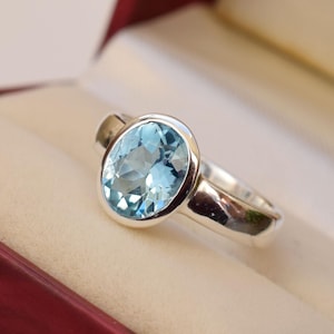 Puede incluir: Un anillo de plata con una piedra ovalada azul claro. El anillo está engastado en una banda simple.