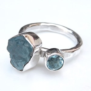 K&ouml;nnte beinhalten: Ein silberner Ring mit zwei blauen Aquamarin-Edelsteinen. Der gr&ouml;&szlig;ere Stein ist ein roher, nat&uuml;rlicher Aquamarin, w&auml;hrend der kleinere Stein ein runder, facettierter Aquamarin ist.